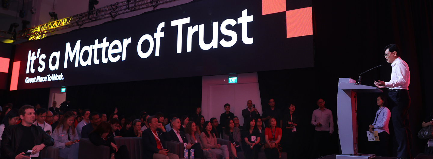 Speaker at podium addresses audience under screen that reads "It's a Matter of Trust."
